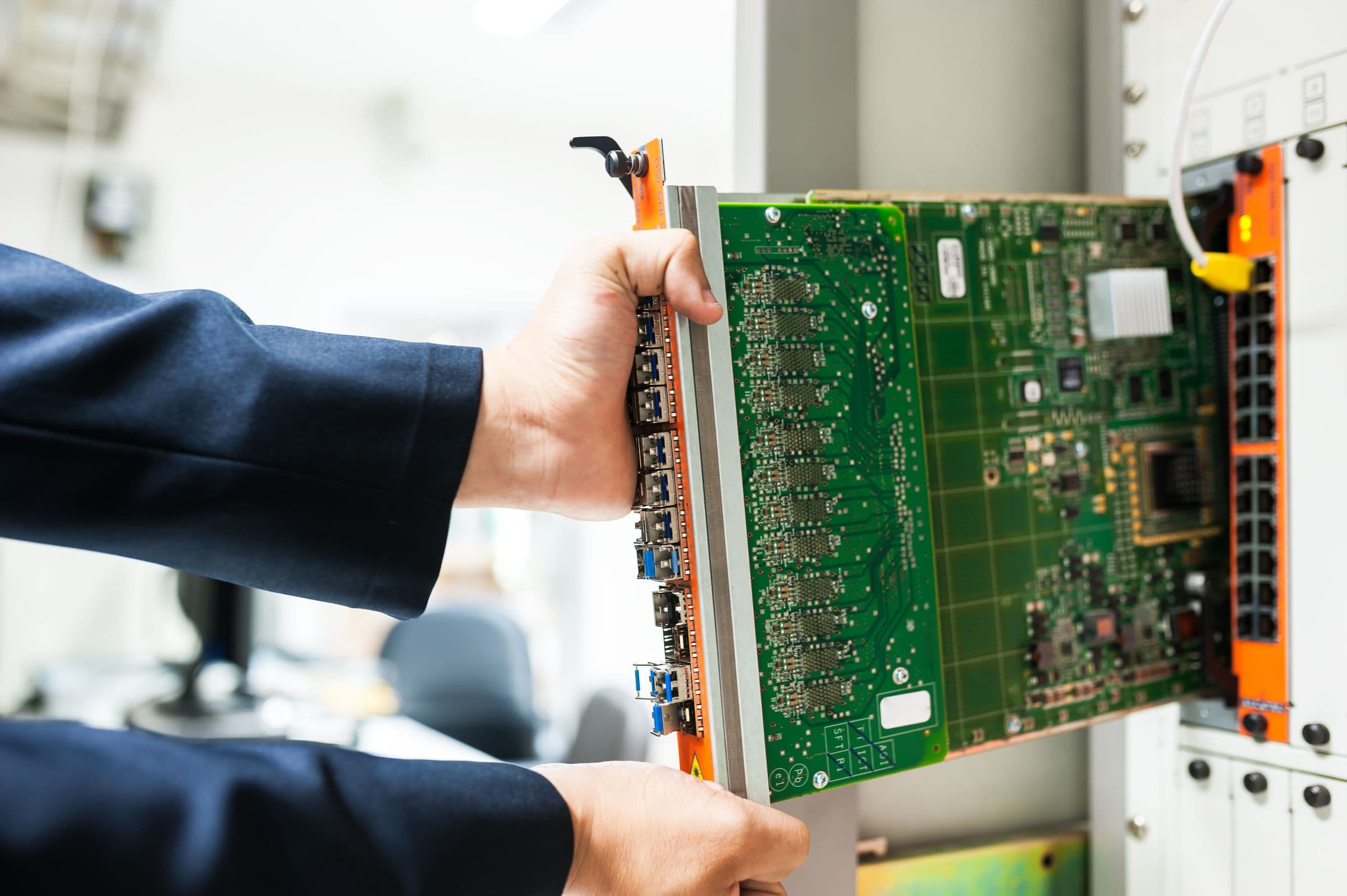 An engineer holding a circuit board beside network equipment.